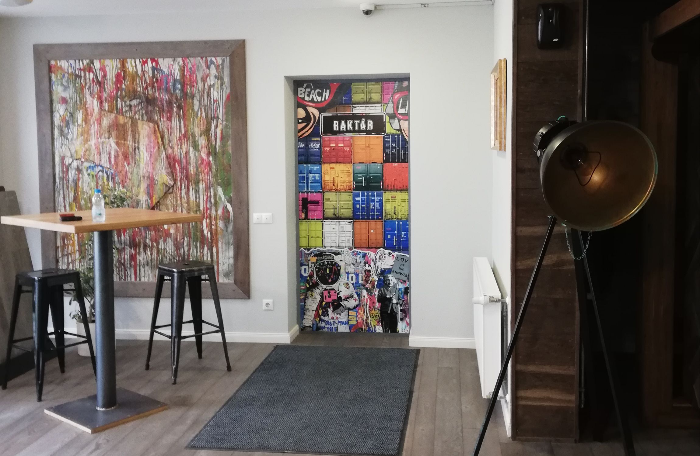 Raktár bejárat, színes konténereket ábrázoló ajtóval, mellette a falon egyedi modern festménnyel